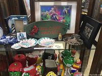 Photo showing a variety of nautical-themed decorative and kitchen items arranged on a glass shelf including shell dishes, a green 'Welcome Aboard' wooden sign, colorful novelty salt and pepper shakers, mugs, and framed art.
