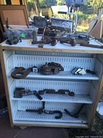 Photo of white metal shelving unit with various large metal hardware placed on shelves and table surface including hooks, pulleys, handles, and rings.