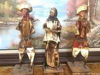 Three corn husk statues representing two men and one woman, standing on rectangular bases, photographed against a natural landscape backdrop.