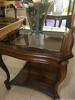 Vintage walnut end table with glass inset top. Curved legs and lower wooden shelf with brass accents visible.