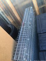 Folded large metal wire panels for an animal crate, stacked with metal hooks along edges, showing parts of black and wooden objects around.