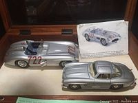 Two detailed Mercedes 300SLR model cars, one open cockpit with racing number 722 and one closed roof coupe with visible details, placed next to a printed information card and a wooden display case.