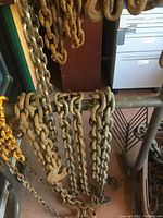 Multiple heavy duty metal chains hanging on pipe rack, showing varied thickness and some with hooks and yellow paint.