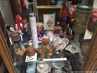 Display shelf with 19 pieces of various collectibles including dolls, wooden items, ceramics, and decorative art, captured in front of a mirror showing reflections.