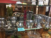 Wide view of 21 pewter items including goblets, bowls, and various decorative pieces on glass shelf.