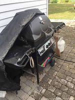 Full view of the Regency 345 Fiesta BBQ grill with a worn cover partially draped over, two propane tanks attached, and utensils hanging from the side shelf.