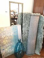 Full lot image showing blue glass vase, floral canvas art, two rolled rugs (one shag, one flat-weave), and a tall ornate framed wall mirror leaning against a wall.