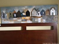 Full view of rectangular canvas wall art showing a row of house shapes in various colors mounted above a wooden panel.