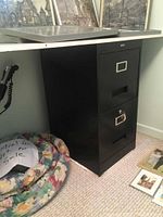 Side view of white pressboard table edge and one black filing cabinet with lock and keys showing.
