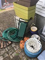 Overview of garden lot items including hose reel, stacked plastic planters, water hose, white plastic plant baskets, blue ceramic planter, and terracotta wall hangings.