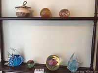 Shelf display showing two blue glass fish figurines, three differently styled glass paperweights including one larger yellow-orange swirl pattern and one with flower and bee embedded inside.