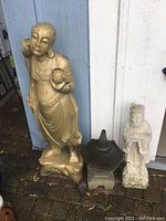 Three Asian inspired lawn ornaments including large beige wood statue, small dark top pagoda-style cement statue, and light colored cement figure statue, all shown with wear