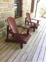 Two maroon painted wood Adirondack chairs flanking a round glass top folding side table on wooden deck.