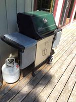 Side view of grill on wooden deck showing green lid, side shelves and attached propane tank