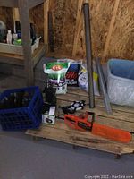 Full view of lot items on wooden pallet in shed floor including electric chainsaw, bags of seed, plastic crate, tire tube, screen, and other items