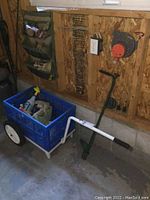 Photo showing metal framed cart with blue plastic crate containing gardening tools next to wall hanging organizer and sprinkler hose reel