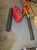 Orange and black Husqvarna gas powered leaf blower with detachable long black plastic blower tube, and large red plastic fuel can with yellow cap on grey floor near wooden pallets.