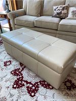 Beige leather storage ottoman positioned in front of a sofa showing overall shape and tufted top