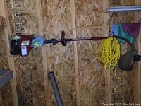 Full view of the red and black gas trimmer hanging on an OSB wall with yellow rope and other items.