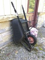 Two images showing black metal frame wheelbarrow with one pink rim wheel and worn condition.