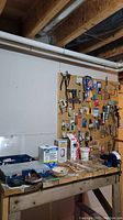 Wide view of wooden workbench against basement wall with pegboard full of hanging tools and the table surface with various hardware boxes and containers, including a metal vice attached to the bench (vice not included in lot).