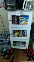 Front view of wood look cabinet with three glass panel doors on three shelves. Cabinet is white and shows chips and scratches. Contents inside are pantry items and soda bottles but are not included.