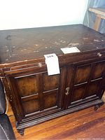 Wooden cabinet with damaged surfaces and two front panel doors, key lock on right door, round knob handles, diamond pattern design on top, standing on small turned legs