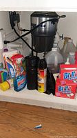 Overview photo showing assortment of cleaning products under a sink including Brillo Basics, Windex, Hotshot, Terro, vinegar, and a white dish rack.