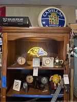 Full view of wooden shelf displaying neon Baltimore Ravens sign lit in yellow, multiple clocks, Cadillac round sign on top, and yellow M&M figure with red object.