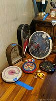 Group of six clocks displayed on wooden table and cabinet, showing various styles, sizes, and visible damage with blue tape tags. Clocks include Joe Boxer flower clock, M&Ms plastic clock, bar-themed 'Tap Room' clock, vintage-carved wooden clock, black clock with red hands, and white circular clock.