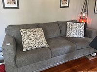 Full view of gray fabric couch with two beige throw pillows having black geometric line patterns. Couch appears in used condition with visible wear. Legs present but couch is on risers.