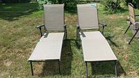 Front view of two beige folding lounge chairs with metal frames on grass. Chairs have armrests and extended leg rests.