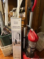 Photo shows boxed Room 2 2-tier rolling garment rack next to a Dirt Devil Endura Reach vacuum in a storage area
