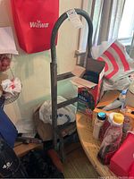 Full view of a metal hand truck standing upright indoors among various household items. Visible curved handle and two horizontal bars for support.