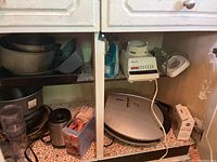 Photo shows kitchen cabinet shelf with George Foreman grill, Farberware blender, Osterizer blender, multiple baking pans, various cooking utensils, and a few glass drinking glasses.