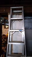 Photo of a 7 ft aluminum ladder with several rungs and cross brace, standing upright against a wall in a basement setting.