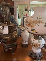Photo showing the floral glass lamp and brass lamp side by side on a table in an indoor room with other furniture and items visible.