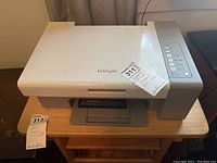 Full view of the Lexmark X2580 printer/scanner unit showing the device on a wooden table with visible labels and dust on the white surface.