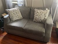 Front view of gray loveseat with two decorative pillows on hardwood floor near window with curtains.