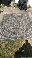 Outdoor rug displayed on concrete and grass. The rug has a concentric circular pattern in shades of gray and off-white. Visible fraying on edges and surface wear can be seen.