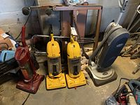 Overview of 4 vacuums: 2 yellow Eureka The Boss Super Lite bagless upright vacuums, 1 red Dirt Devil Dynamite vacuum, and 1 silver/gray Halo upright vacuum all on concrete floor in basement area, showing wear and dust.