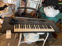 Front view of Yamaha PSR-27 keyboard placed on white plastic stand in a cluttered basement environment, showing full keyboard and control panel with power cord attached.