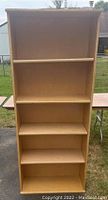Full view of the wooden 5-shelf bookcase standing outside on grass with backdrop showing some wear and missing pins.