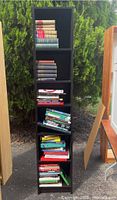Tall black bookshelf holding approximately 40 educational books, organized in five compartments positioned outdoors on a paved surface.