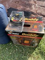 Photo showing Mr. Heater Portable Buddy heater box, cords for larger propane tanks, and a blue sleeping bag rolled up next to it, outside on grass.