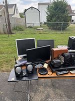 Wide view of table with multiple monitors, speakers, vintage cameras, audio equipment, and accessories arranged outdoors on asphalt