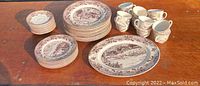 Full view of multiple plates, serving tray, and coffee mugs arranged on wooden surface showing the scenic brown transferware design.