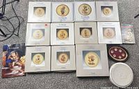 Photo showing 11 boxed Hummel plates arranged including the framed mini plate collection and two collector guides, plus an Easter plate and anniversary plate box