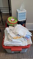 Photo showing stacked tins and tissue box cover placed on top of a large plastic storage bin filled with folded white linens.