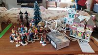 Group of miniature Christmas village figurines including people, two LED street lamps, a green bottle brush Christmas tree, two buildings resembling a hospital and railway station, with electrical cords and a matchbox for scale.
